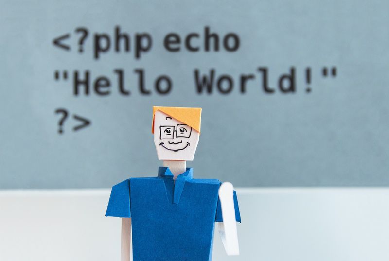 Deko-Bild: Grüßende Papierfigur vor einem im Hintergrund leicht verschwommen erscheinenden Code, der in PHP »Hello World« ausgibt.